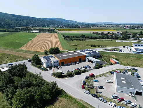 Luftaufnahme eines Firmengebäudes mit orangefarbener Fassade, umgeben von Feldern und Hügeln, mehrere Autos auf dem Parkplatz.