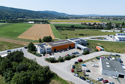 Luftaufnahme eines Firmengebäudes mit orangefarbener Fassade, umgeben von Feldern und Hügeln, mehrere Autos auf dem Parkplatz.