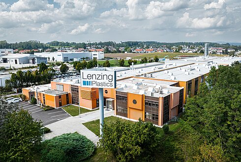 Luftaufnahme einer Industrieanlage, mit orangefarbenen Fassadenelementen, weißem Dach und umliegenden Grünflächen. Im Vordergrund ein großes Schild „Lenzing Plastics