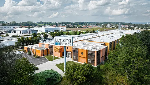Luftaufnahme einer Industrieanlage, mit orangefarbenen Fassadenelementen, weißem Dach und umliegenden Grünflächen. Im Vordergrund ein großes Schild „Lenzing Plastics