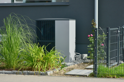 Eine Wärmepumpe steht in einem Blumenbeet. Sie wird teilweise von hohem Gras verdeckt.