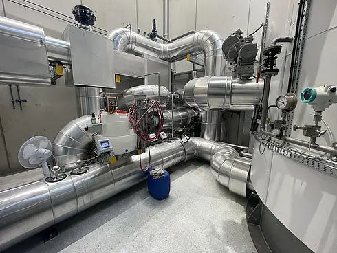 Ein komplexes industrielles Setup mit zahlreichen miteinander verbundenen Edelstahlrohren und Maschinen. Sichtbar sind verschiedene Messgeräte, Ventile und Steuerpulte. In der Bildmitte steht ein blauer Fass auf dem Boden. Die Umgebung wirkt sauber und gut gepflegt, vermutlich Teil einer Verarbeitungs- oder Produktionsanlage.