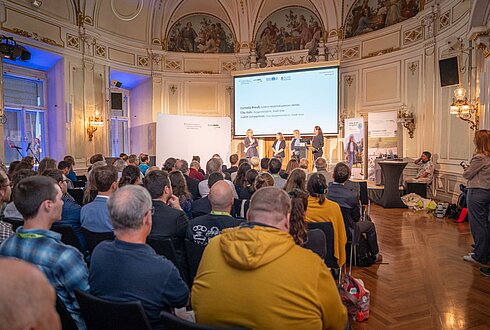 Prunkvoller Saal mit vielen Menschen, die sitzend lauschen