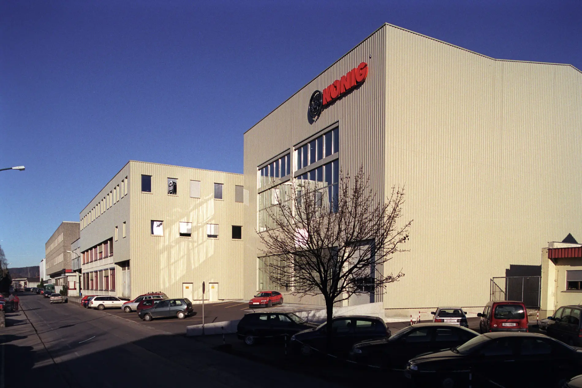 Außnansicht der König GmbH & Co KG in Rankweil. Auf der Fassade spiegelt sich das Sonnenlicht unter strahlend blauem Himmel. Vor den Werkshallen stehen parkende Fahrzeuge und ein Baum.