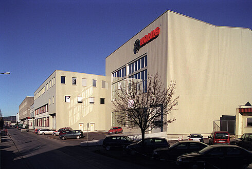 Außnansicht der König GmbH & Co KG in Rankweil. Auf der Fassade spiegelt sich das Sonnenlicht unter strahlend blauem Himmel. Vor den Werkshallen stehen parkende Fahrzeuge und ein Baum.