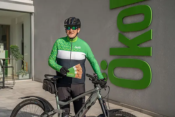 Mann mit Helm und ÖkoFEN Fahrradtrikot, steht hinter Fahrrad, hält Frühstücksgutscheine in der Hand