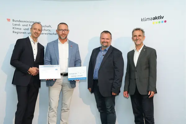 Menschen mit einer Urkunde in der Hand vor einer Fotowand. Sie lächeln in die Kamera.