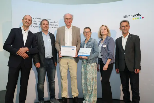 Menschen mit einer Urkunde in der Hand vor einer Fotowand. Sie lächeln in die Kamera.