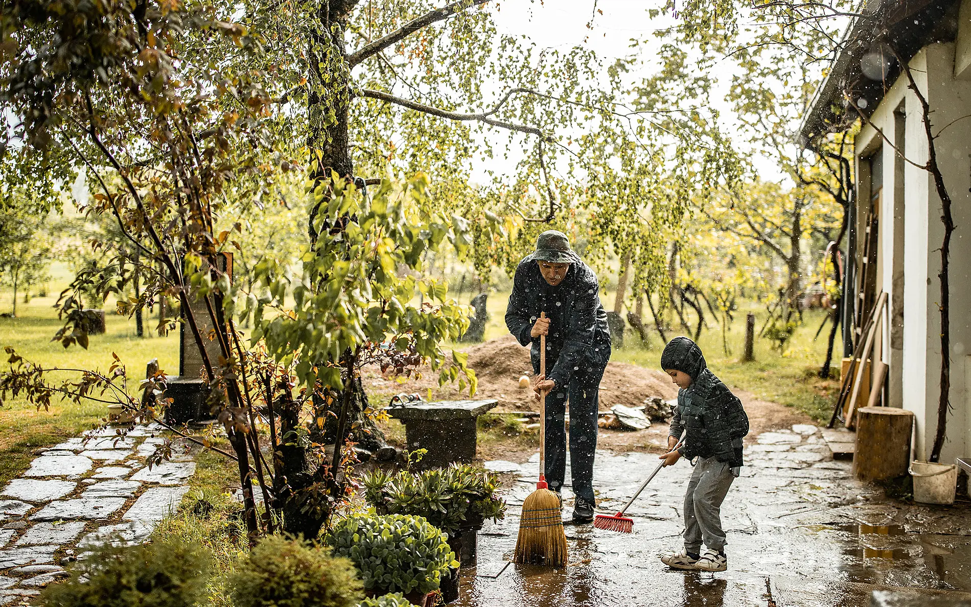 Eine erwachsene Person und ein Kind kehren nach einem starken Regen Wasser von einer gepflasterten Fläche vor einem Haus. Der Garten und die Wege sind nass, Regen fällt durch die Bäume.