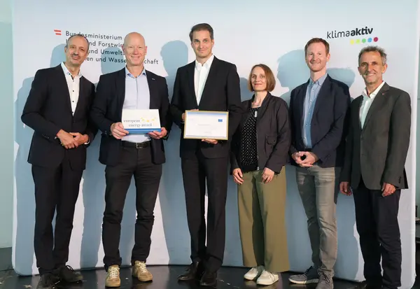 Menschen mit einer Urkunde in der Hand vor einer Fotowand. Sie lächeln in die Kamera.
