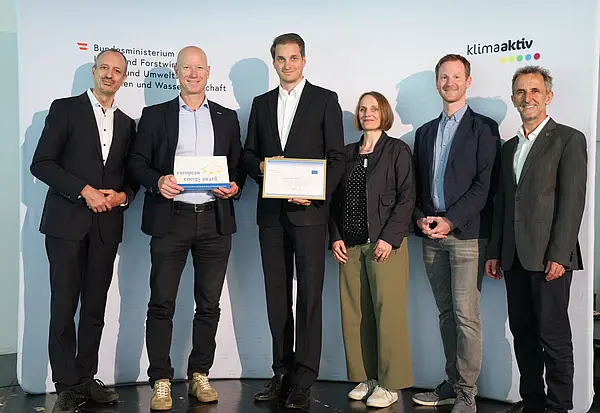 Menschen mit einer Urkunde in der Hand vor einer Fotowand. Sie lächeln in die Kamera.