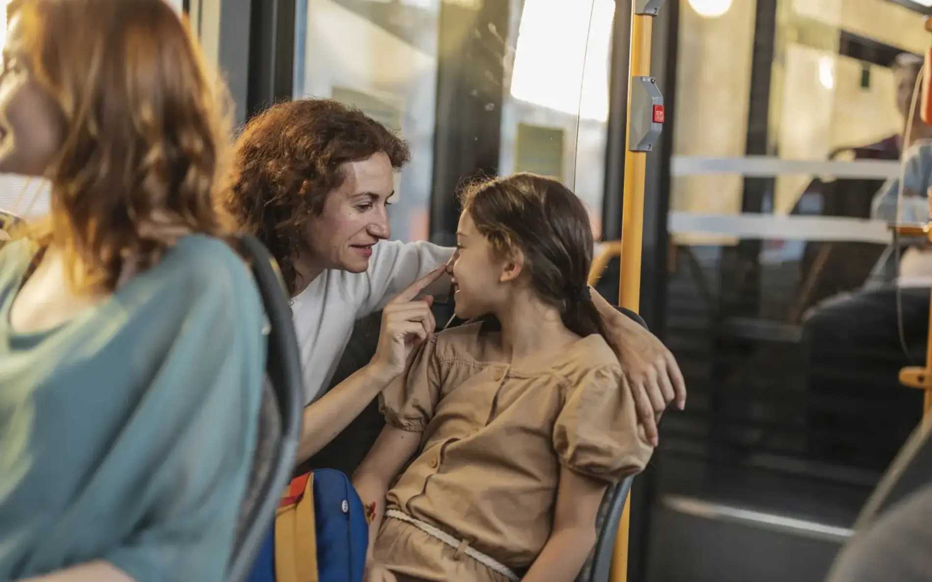 Mutter und Tochter sitzen nebeneinander im Bus, die Mutter streift spielerisch über ihre Nase.