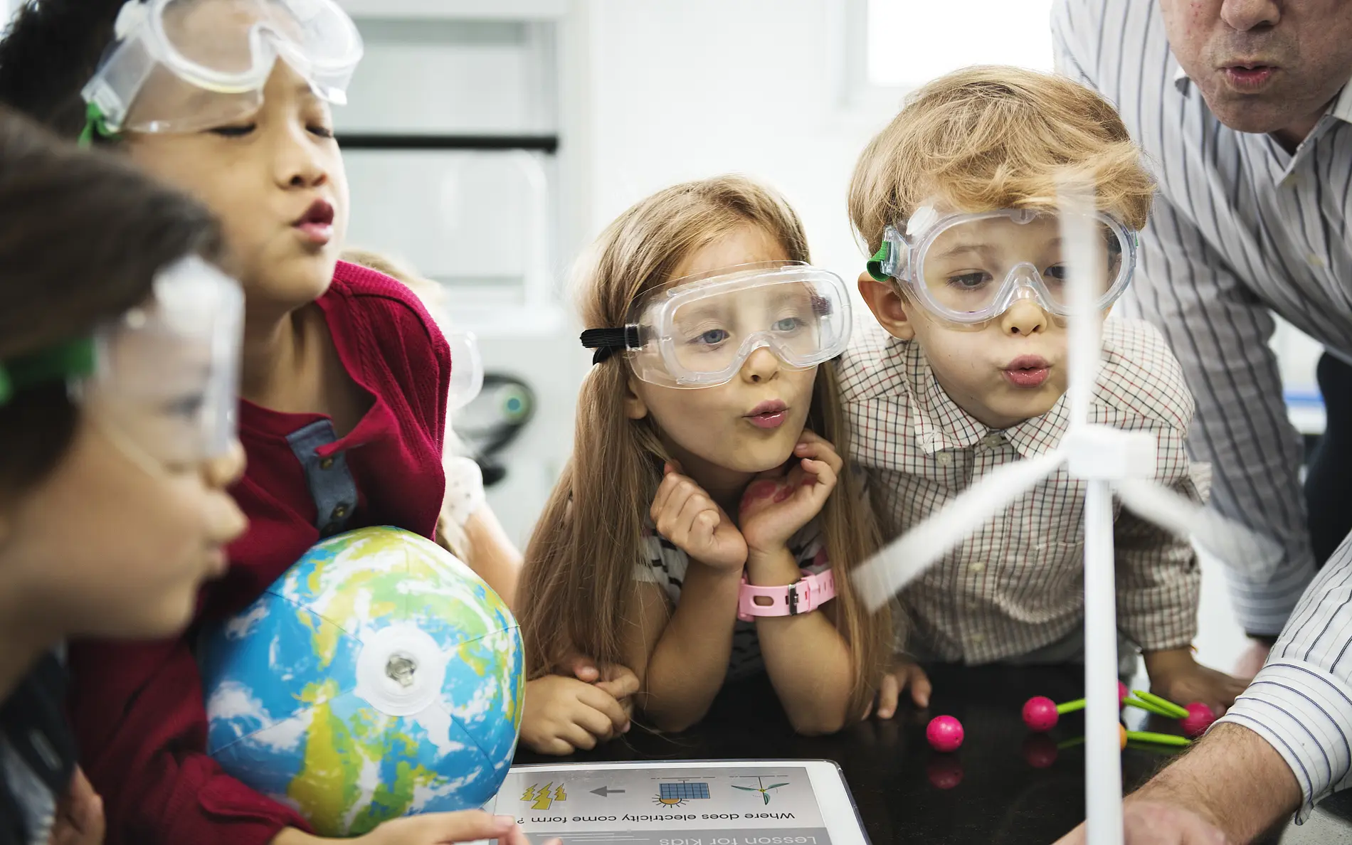 Mehrere Kinder tragen Schutzbrillen und stehen gemeinsam an einem Tisch in einem Unterrichts- oder Experimentierraum. Im Vordergrund befindet sich ein Modell einer Windkraftanlage, dessen Rotorblätter von den Kindern durch Pusten in Bewegung versetzt werden. Ein weiteres erwachsenes Gegenüber beugt sich von der rechten Bildseite zum Experimentiertisch.  Auf dem Tisch liegen Lernmaterialien zum Thema erneuerbare Energien, darunter eine Tafel mit Abbildungen von Windkraft- und Solaranlagen. Außerdem ist ein Globus zu sehen. Die Kinder konzentrieren sich auf das Windradmodell und beobachten dessen Reaktion.  Der Raum ist hell ausgeleuchtet und wirkt wie ein naturwissenschaftlicher Unterrichts- oder Workshopraum. Das Bild vermittelt spielerisches Lernen und Experimentieren zu den Themen Energie, Technik und Nachhaltigkeit.