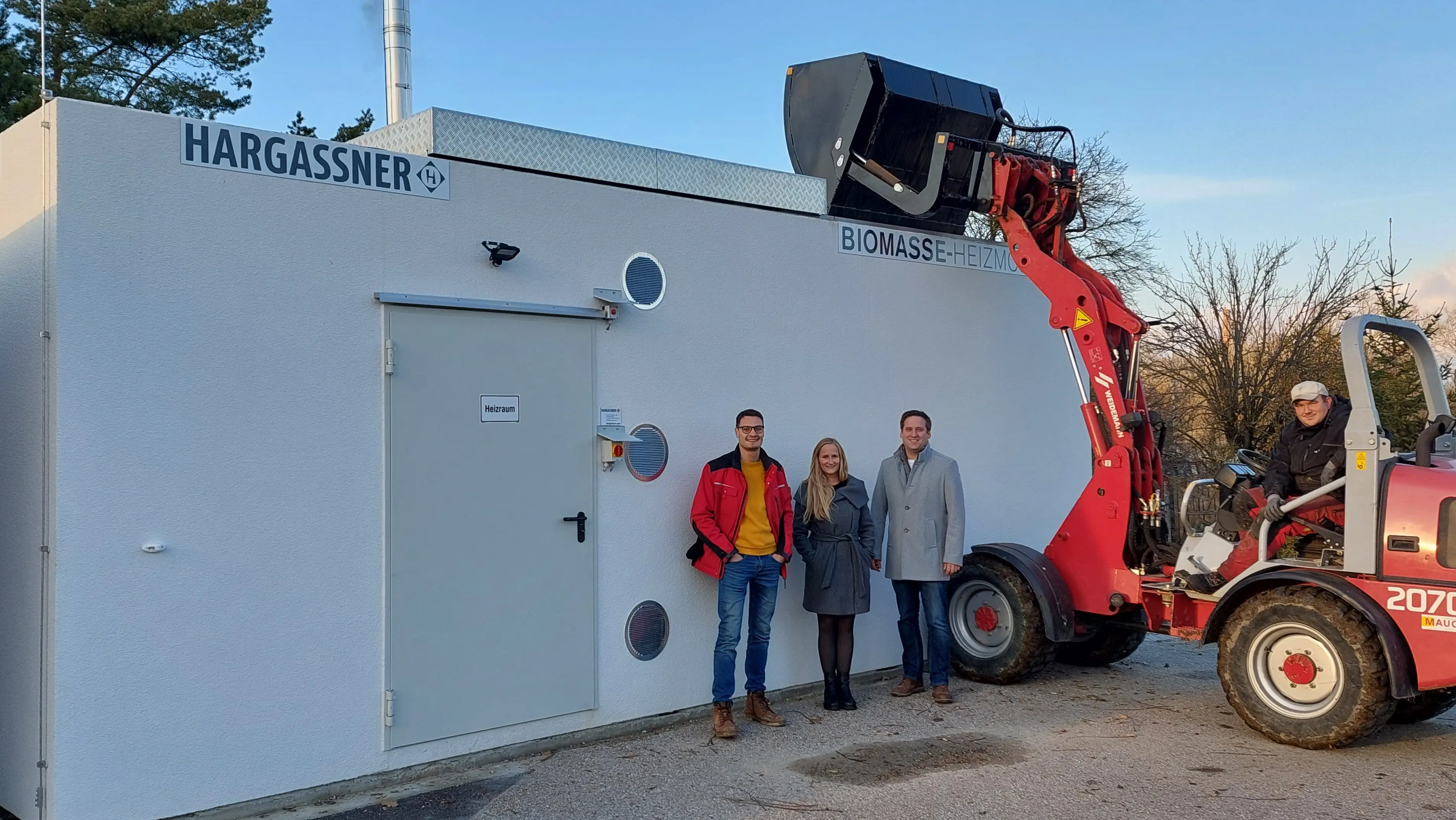 Drei Personen stehen vor einem weißen Gebäude mit der Aufschrift „Hargassner Biomasse-Heizzentrale“, daneben ein roter Radlader.