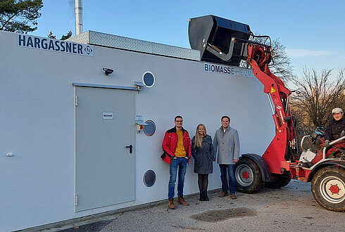 Drei Personen stehen vor einem weißen Gebäude mit der Aufschrift „Hargassner Biomasse-Heizzentrale“, daneben ein roter Radlader.