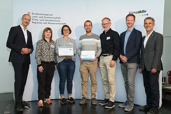 Menschen mit einer Urkunde in der Hand vor einer Fotowand. Sie lächeln in die Kamera.