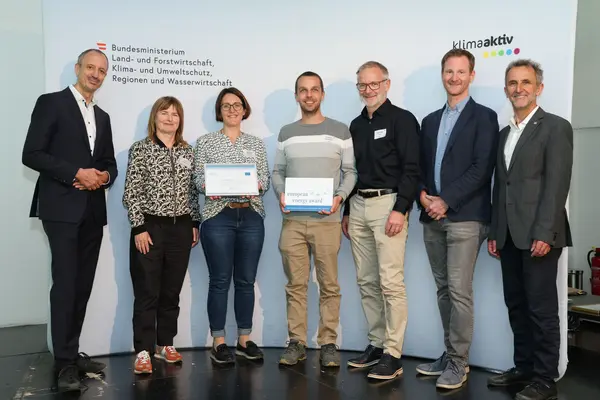 Menschen mit einer Urkunde in der Hand vor einer Fotowand. Sie lächeln in die Kamera.