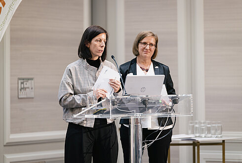 Zwei Personen hinter einem Rednerpult mit Mikrofon und Laptop, daneben ein Tisch mit Wassergläsern.