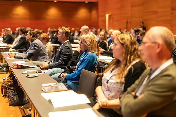 Mehrere Personen sitzen an einem langen Tisch und lauschen der Konferenz