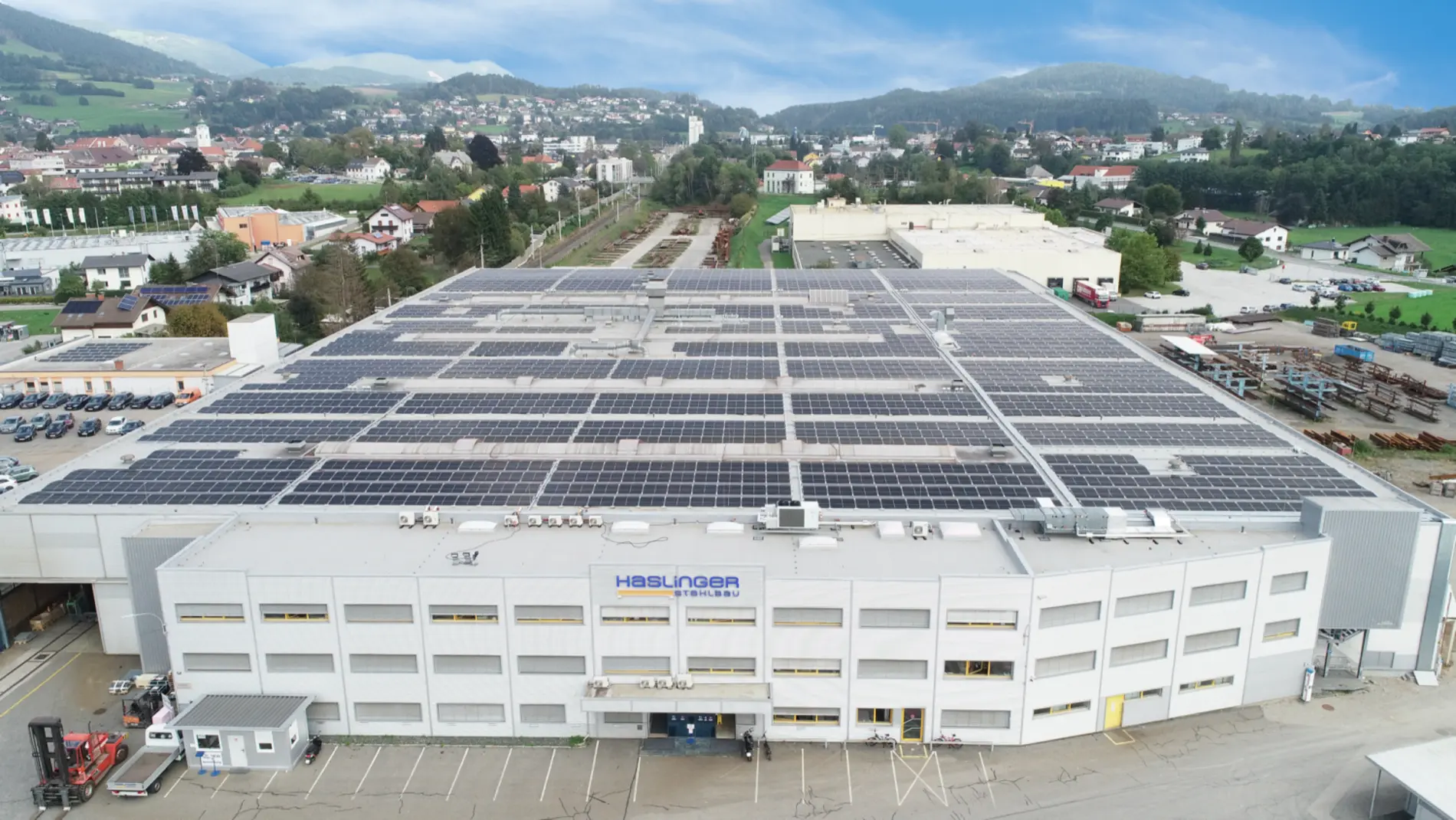 Unternehmensbild der Haslinger Stahlbau GmbH. Vogelperspektive auf eine große Halle mit einer gebirgigen Kulisse unter leicht bewölktem Himmel im Hintergrund. Auf dem Dach sind flächendeckend zahlreiche PV-Paneele installiert.