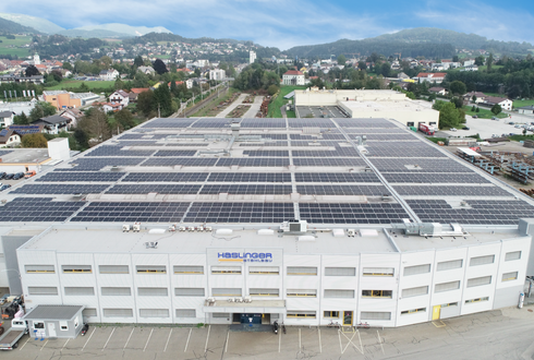 Unternehmensbild der Haslinger Stahlbau GmbH. Vogelperspektive auf eine große Halle mit einer gebirgigen Kulisse unter leicht bewölktem Himmel im Hintergrund. Auf dem Dach sind flächendeckend zahlreiche PV-Paneele installiert.