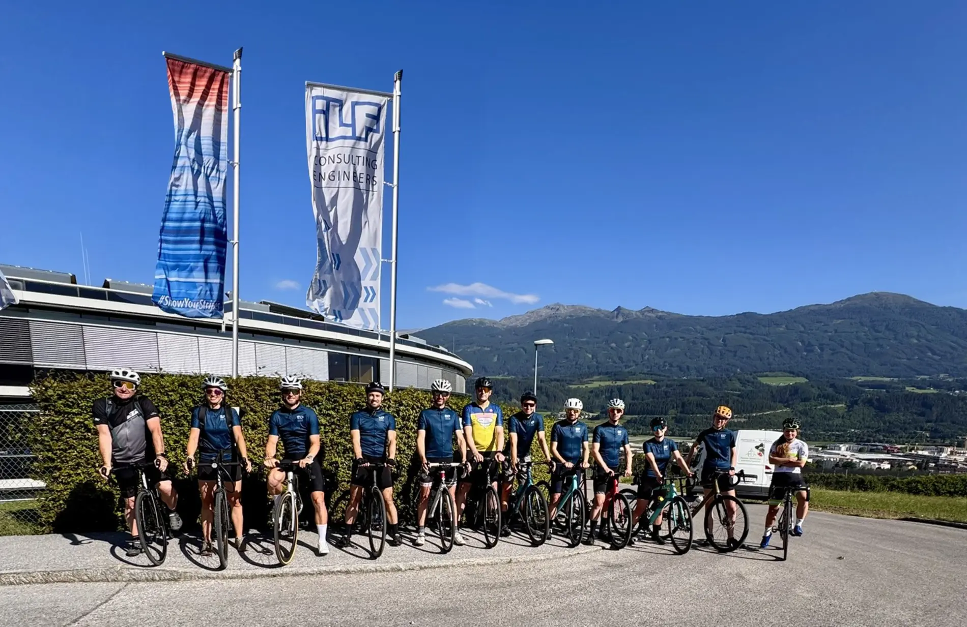 Viele Radfahrer:innen stehen nebeneinander mit gleichem Fahrradtrikot auf der Straße, dahinter wehen zwei Fahnen im Wind