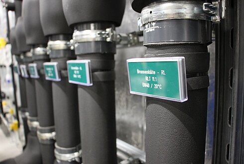 Mehrere isolierte Rohre mit Etiketten, die technische Angaben enthalten. Eine sichtbare Beschriftung lautet: „Brunnenkälte – RL, RLT 11.1, DN40 / 20°C“.