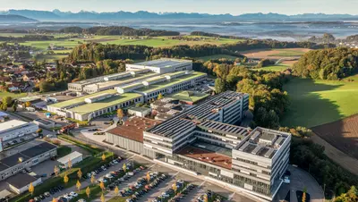 Luftaufnahme des Werksgeländes der B&R Automation GmbH am Standort Eggelsberg. Mehrere Gebäude und ein großer Parkplatz vor einer idyllischen Landschaft und malerischen Bergkulisse.