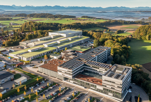 Luftaufnahme des Werksgeländes der B&R Automation GmbH am Standort Eggelsberg. Mehrere Gebäude und ein großer Parkplatz vor einer idyllischen Landschaft und malerischen Bergkulisse.