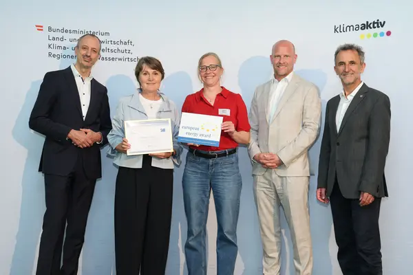 Menschen mit einer Urkunde in der Hand vor einer Fotowand. Sie lächeln in die Kamera.