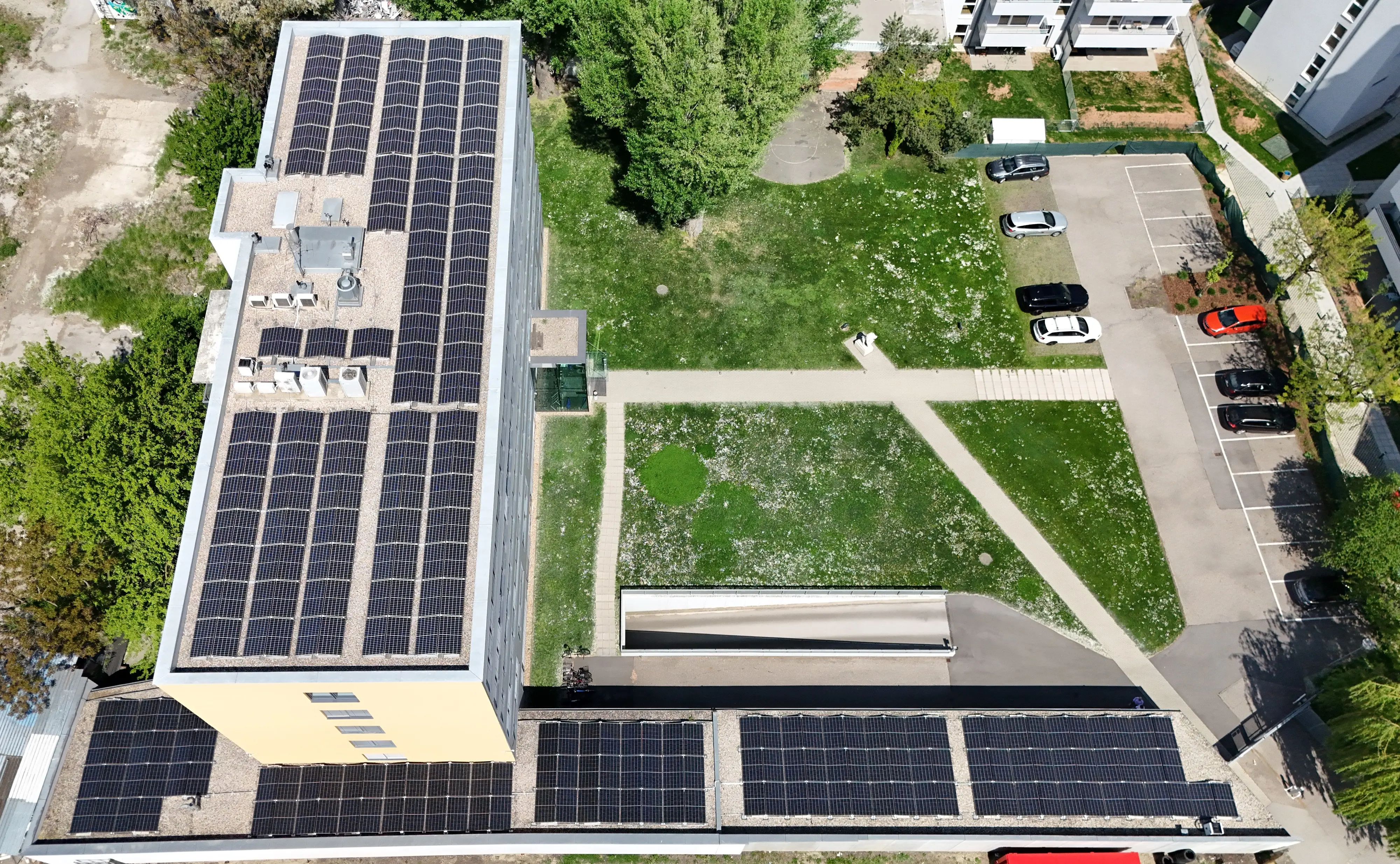 Luftaufnahme eines Gebäudedachs mit Solarpaneelen, angrenzend an eine grüne Wiese und einen Parkplatz.
