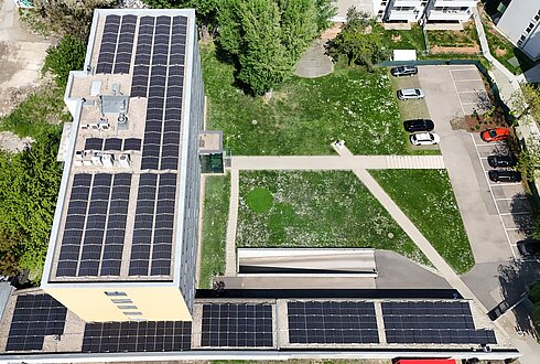 Luftaufnahme eines Gebäudedachs mit Solarpaneelen, angrenzend an eine grüne Wiese und einen Parkplatz.