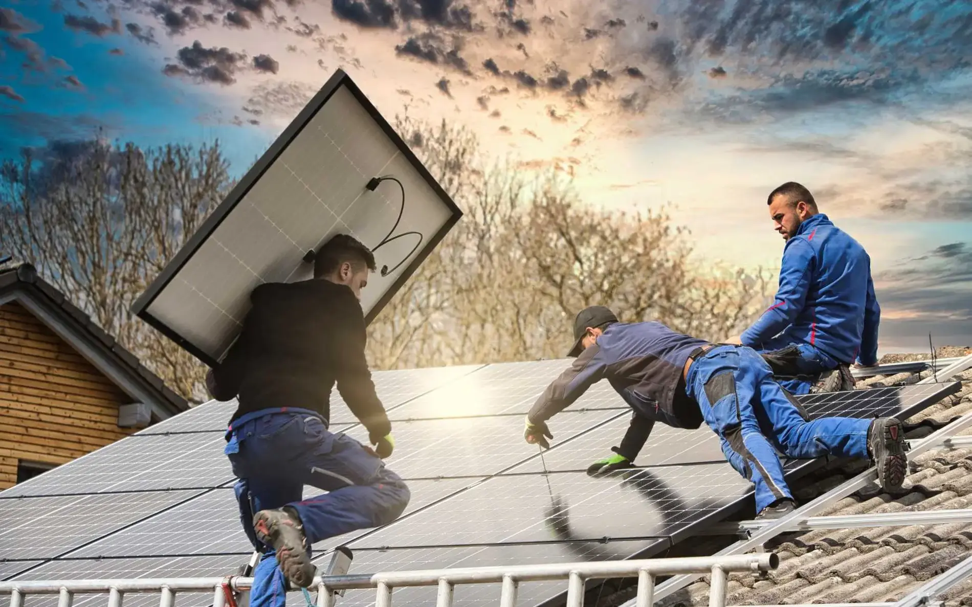 Drei Männer befestigen Solarpanele am Dach. Die Hälfte der Photovoltaikanlage ist bereits fertig. Einer der Männer klettert gerade mit einem Panel auf der Schulter die Leiter hoch.