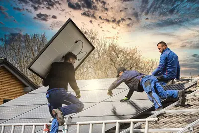 Drei Männer befestigen Solarpanele am Dach. Die Hälfte der Photovoltaikanlage ist bereits fertig. Einer der Männer klettert gerade mit einem Panel auf der Schulter die Leiter hoch.