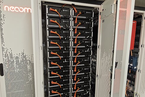 Geöffneter Batteriespeicher-Schrank mit mehreren schwarzen Batteriemodulen und orangefarbenen Kabeln, Teil eines Energiespeichersystems.