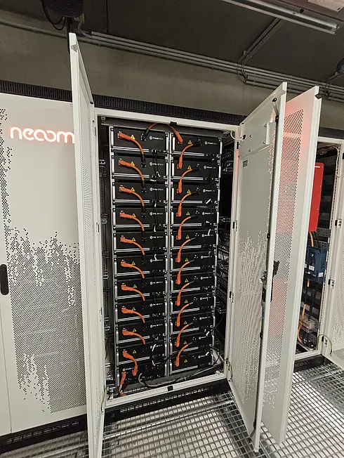 Geöffneter Batteriespeicher-Schrank mit mehreren schwarzen Batteriemodulen und orangefarbenen Kabeln, Teil eines Energiespeichersystems.