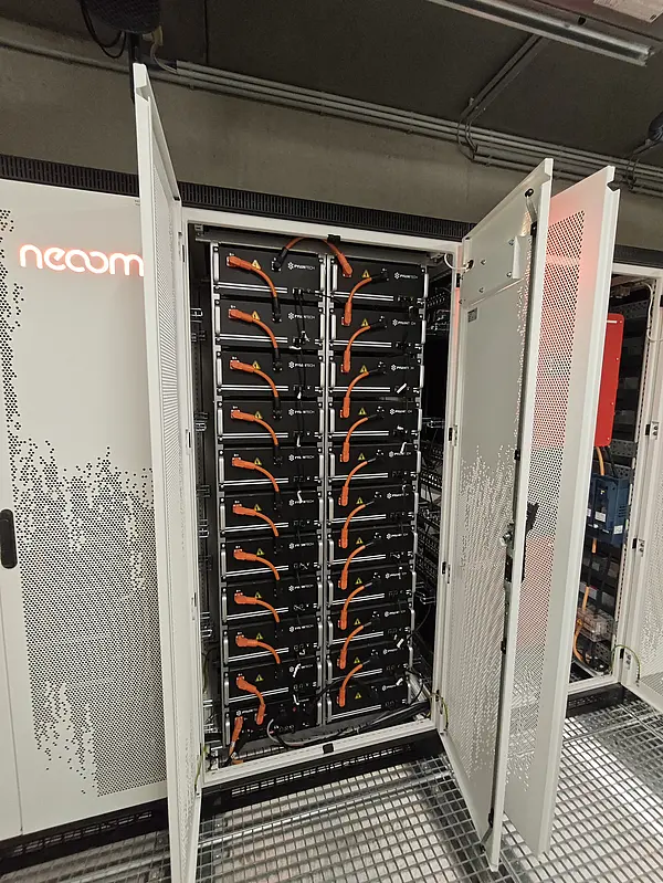 Geöffneter Batteriespeicher-Schrank mit mehreren schwarzen Batteriemodulen und orangefarbenen Kabeln, Teil eines Energiespeichersystems.
