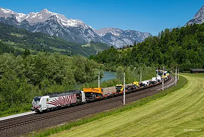 Güterzug fährt durch grüne Landschaft zwischen Feld und Fluss, Berge im Hintergrund