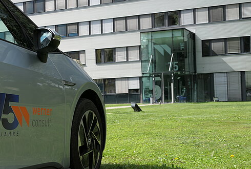 Ein Auto mit dem 75-Jahre-Jubiläumslogo von Werner Consult steht vor dem Eingang des Bürogebäudes mit der Hausnummer 15.