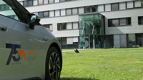 Ein Auto mit dem 75-Jahre-Jubiläumslogo von Werner Consult steht vor dem Eingang des Bürogebäudes mit der Hausnummer 15.