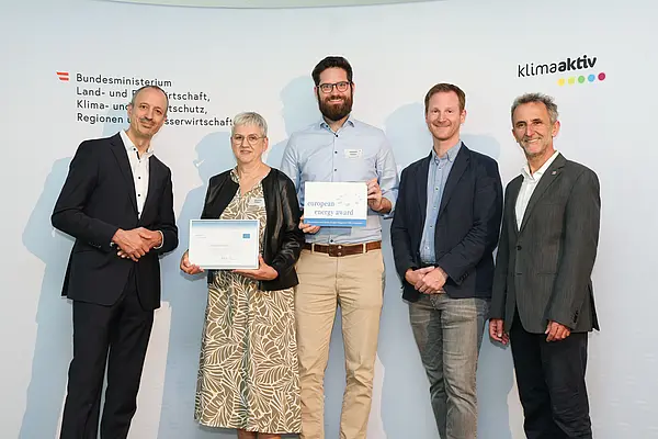 Menschen mit einer Urkunde in der Hand vor einer Fotowand. Sie lächeln in die Kamera.