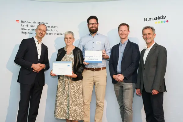 Menschen mit einer Urkunde in der Hand vor einer Fotowand. Sie lächeln in die Kamera.