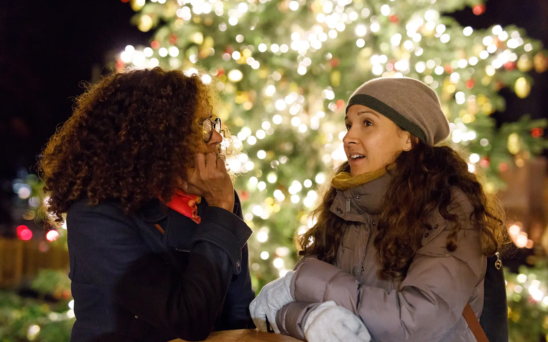 Zwei Frauen stehen vor einem beleuchteten Weihnachtsbaum im Freien und sprechen miteinander.