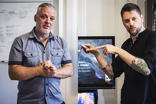Michael Kellermann (links) und Philip Herberstein (rechts) stehen in einem Innenraum neben einem Bildschirm. Kellermann trägt ein kurzärmeliges Hemd und hält die Hände vor dem Körper. Herberstein hält eine Fernbedienung in der Hand und zeigt mit dem Finger darauf. Im Hintergrund sind ein Whiteboard sowie ein Bildschirm mit teilweise sichtbarer Präsentationsfolie zu sehen.