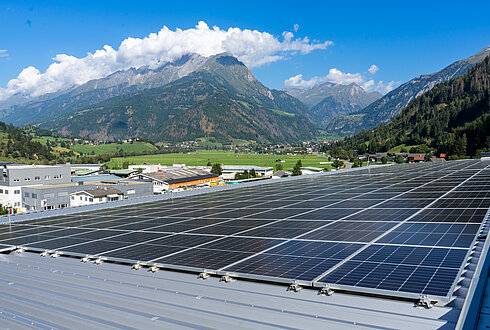 Solarpaneele auf einem geneigten Metalldach mit Blick auf Gebäude, Felder und Berge im Hintergrund.