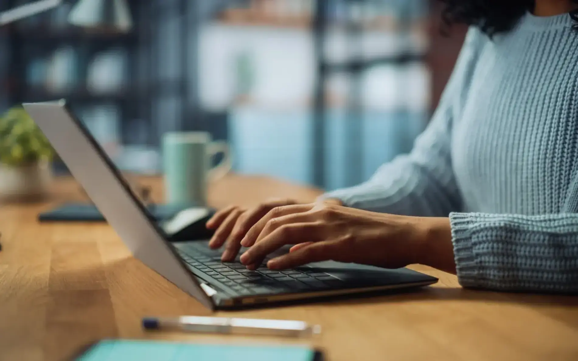 Ein Laptop steht am Holztisch, Frauenhände tippen gerade auf der Tastatur. Rechts der Person steht eine Tasse, links davon ein Smartphone, dessen Bildschirm an ist.