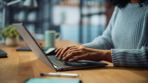 Ein Laptop steht am Holztisch, Frauenhände tippen gerade auf der Tastatur. Rechts der Person steht eine Tasse, links davon ein Smartphone, dessen Bildschirm an ist.