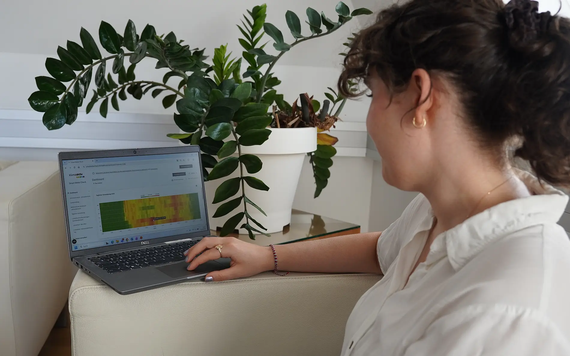 Eine Person sitzt in einem hellen Wohn- oder Arbeitsraum auf einem Sofa und nutzt einen Laptop. Auf dem Bildschirm ist ein Dashboard mit einer farbcodierten Grafik in Grün-, Gelb- und Rottönen zu sehen. Die Person bedient das Touchpad und blickt auf die dargestellten Daten.  Neben dem Laptop steht eine große Zimmerpflanze in einem weißen Topf auf einem Beistelltisch. Der Raum ist modern und hell eingerichtet, mit weißen Wänden und Möbeln. Tageslicht fällt von rechts in den Raum und sorgt für eine freundliche Atmosphäre.  Das Bild zeigt die digitale Auswertung von Energie- oder Verbrauchsdaten im Wohnumfeld und veranschaulicht die Nutzung eines Online-Tools zur Analyse und Optimierung des Energieverbrauchs.