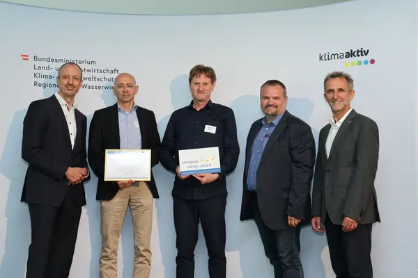 Menschen mit einer Urkunde in der Hand vor einer Fotowand. Sie lächeln in die Kamera.