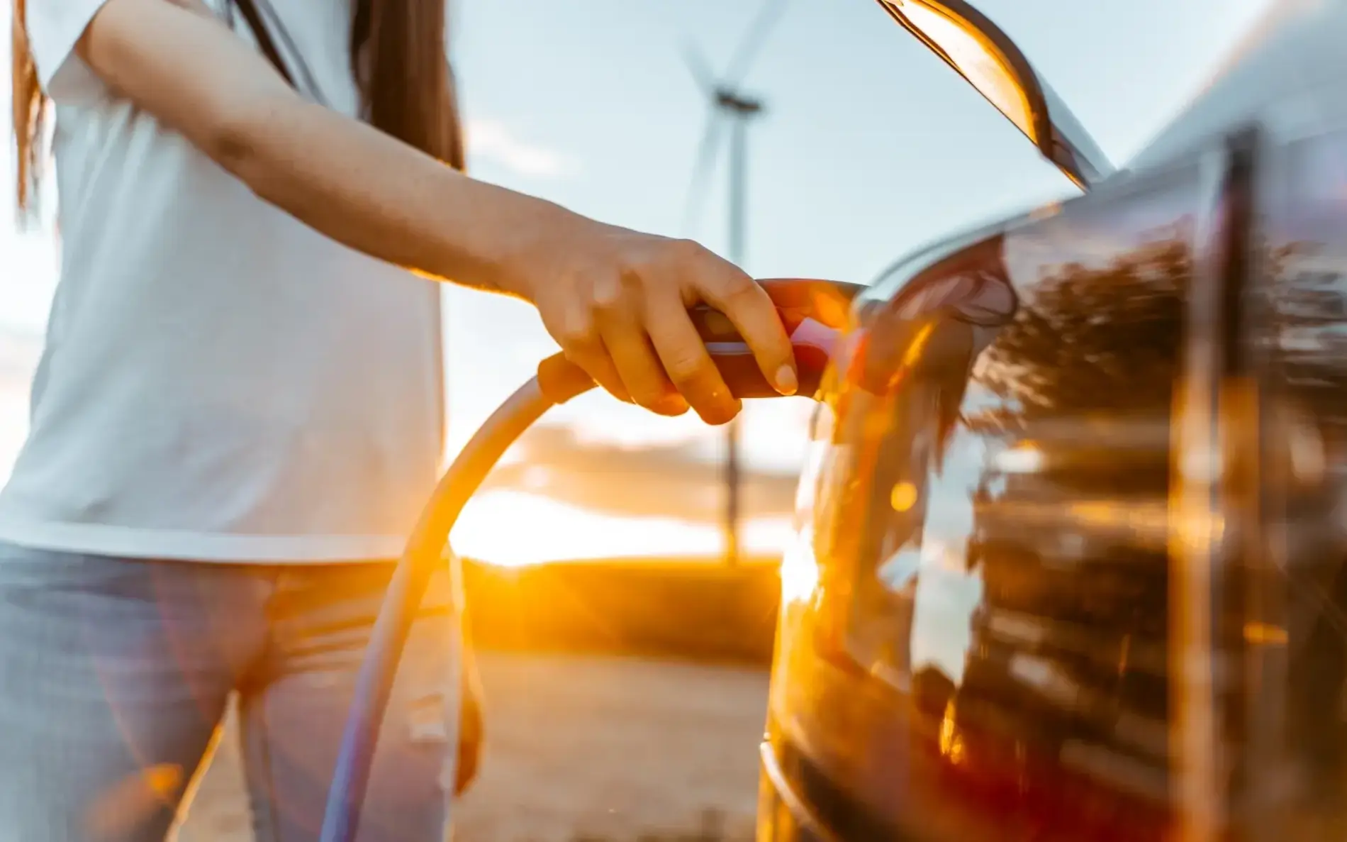 Ein Mann steht im SOnnenuntergang, im Hintergrund ist ein Windrad zu sehen, er hat dabei ein Ladkabel in der Hand, was er in sein E-Auto steckt. 